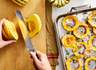 A pair of hands slices a delicata squash on a wooden cutting board, with an 8 inch Misen Serrated Knife. The slices are then arranged in a Misen Roasting Pan alongside slices of red onion and herbs.