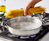 A hand holds a large bundle of dry spaghetti noodles over a Misen stainless steel rondeau of actively boiling water on a gas stove. A blue flame is visible beneath the pan.