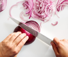 A pair of hands is shown slicing a red onion on a white cutting board using a Misen knife. Thinly sliced pieces of red onion are scattered around the work area.