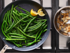 An overhead shot of a Misen Carbon Nonstick™ Frying Pan on a gas stovetop, filled with sauteed green beans seasoned with salt. Two lemon wedges are resting on the upper right side of the pan. To the right of the pan, a smaller stainless steel saucier contains sliced almonds and a chef spoon.