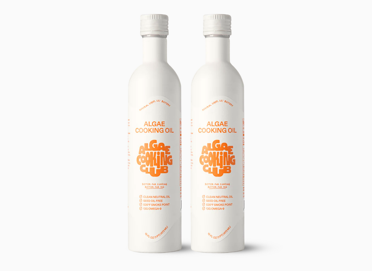 Two white bottles of Algae Cooking Oil are shown, each with a white cap. The front of each bottle features the text "ALGAE COOKING OIL" in orange, above a stylized "ALGAE COOKING CLUB" logo in orange and yellow. Below the logo is the text "BETTER FOR COOKING. BETTER FOR YOU." in smaller red text. Further down, in small orange text, is a list of features: "CLEAN NEUTRAL OIL," "SEED OIL FREE," "535°F SMOKE POINT," and "13G OMEGA-9." The bottom of each bottle is marked "16 FL OZ (1 PT) (473 ML)."