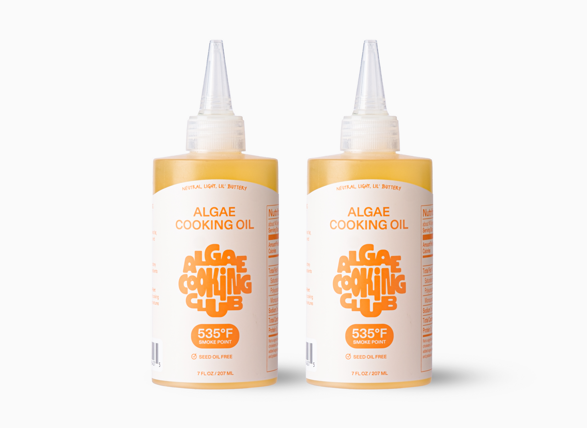 Two clear bottles of "Algae Cooking Oil" by the Algae Cooking Club are depicted in the image. Each bottle has an orange and white label that states "535°F Smoke Point" and "Seed Oil Free." The bottles each contain 7 FL OZ / 207 ML of oil.