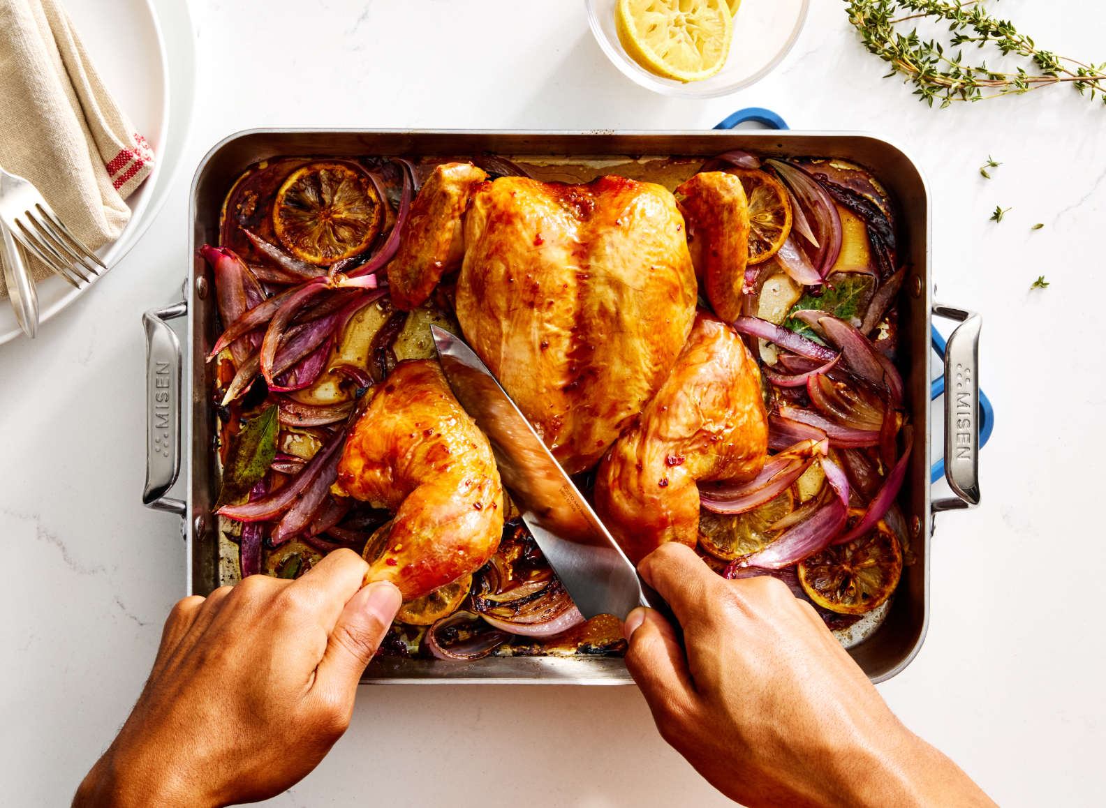 A top-down view shows a person carving a roasted chicken in a stainless steel roasting pan. The chicken is surrounded by roasted red onions and lemon slices. The roasting pan has "MISEN" embossed on its handles, and it sits on a white countertop next to a small bowl of lemon wedges and a sprig of thyme.