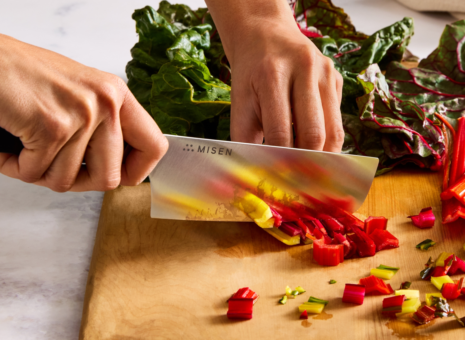 The image shows a close-up of a person's hands holding a large, rectangular Nakiri Knife with the word "MISEN" visible on the blade. The person is using the knife to chop colorful Swiss chard stalks and leaves on a light wooden cutting board.