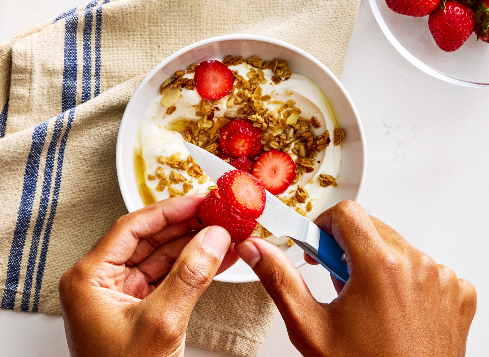 A top-down view shows a person's hands holding a red strawberry and a small, Misen ACUTO440 3.5 inch paring knife with a blue handle over a white bowl of yogurt, granola, and sliced strawberries. The knife is positioned to slice the strawberry. A blue striped Misen kitchen towel is draped over the top left corner of the bowl, and a clear bowl filled with whole strawberries is visible in the upper right corner of the image.