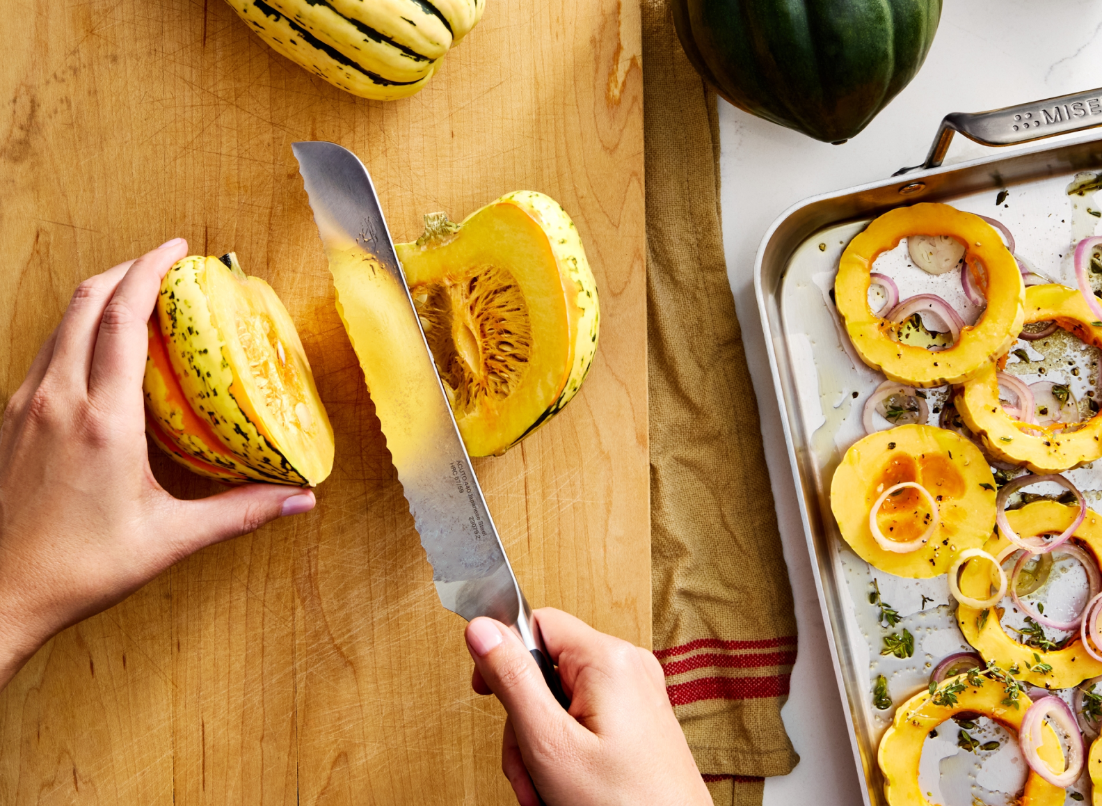A top-down view shows a person slicing a delicata squash with an 8 inch ACUTO440 Misen serrated knife on a wooden cutting board. The squash is yellow with green stripes, and one half of it, with seeds exposed, sits to the right of the knife. The knife has "MISEN" engraved on its blade. To the right of the cutting board, a Misen 18-inch stainless steel roasting pan filled with roasted delicata squash rings, red onion, and herbs is visible. A dark green acorn squash is in the upper right corner of the image.