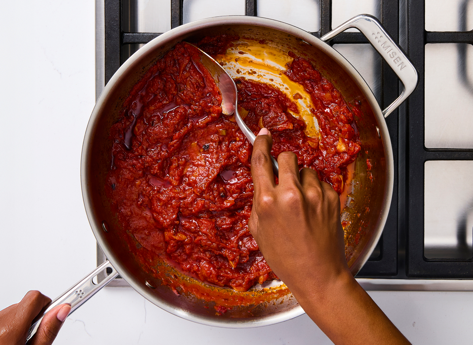 An overhead shot shows a 4 qt stainless steel all-purpose pan on a stovetop, filled with a thick red sauce that a hand is stirring with a chef spoon. The handle on the right side of the pan has "MISEN" inscribed on it.