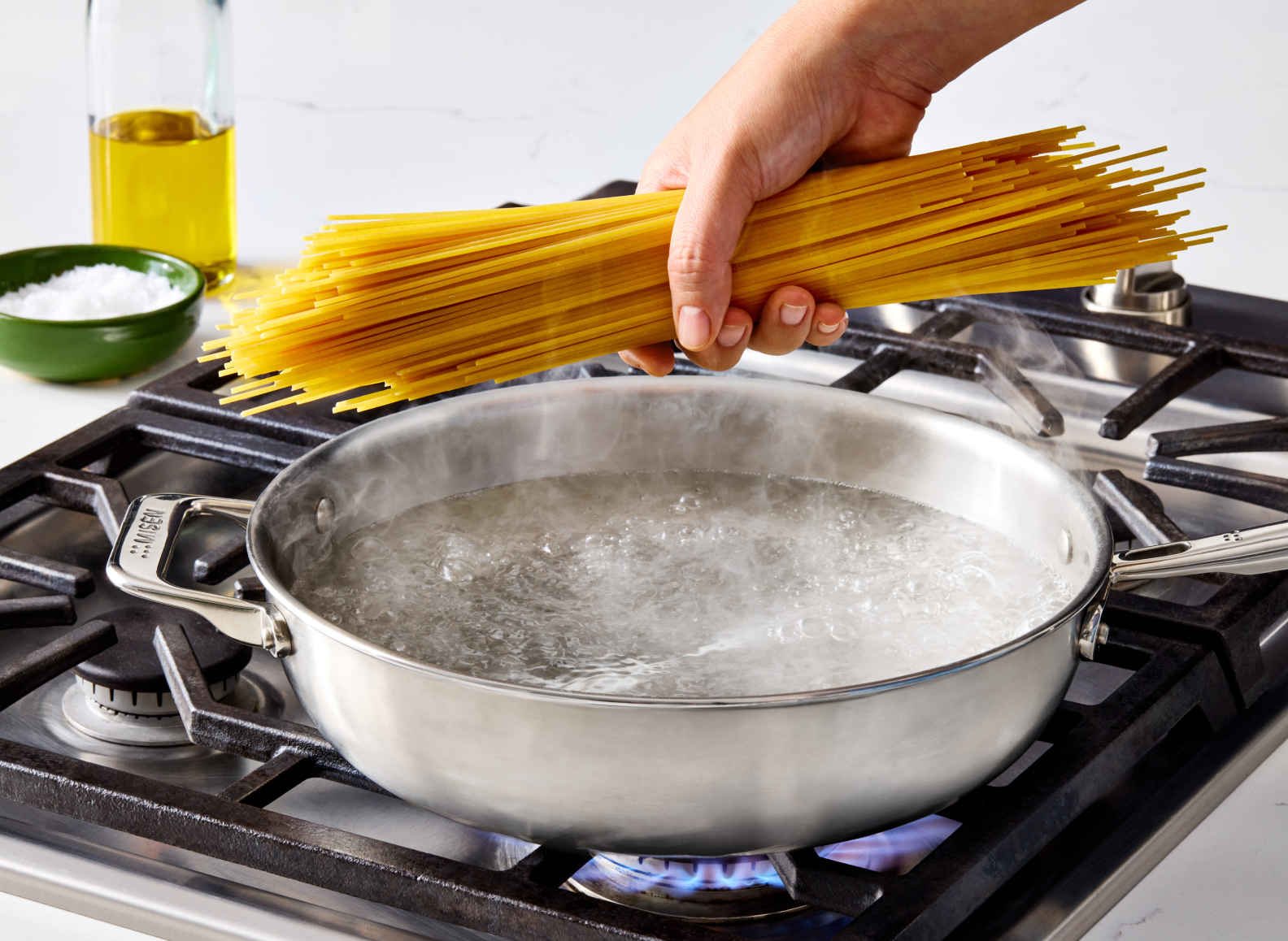 A hand is shown holding a bundle of dry spaghetti noodles over a 4 QT stainless steel all-purpose pan of boiling water on a gas stovetop. To the left on the countertop, there is a bottle of olive oil and a small bowl of salt. The pan has a handle on the left side with "MISEN" embossed on it.