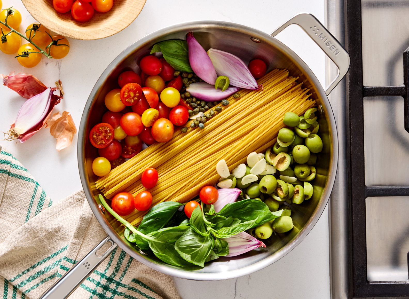 An overhead shot of a 4 QT stainless steel all-purpose pan on a stovetop shows dry spaghetti noodles surrounded by various raw ingredients for a pasta dish, including whole red and yellow cherry tomatoes, green olives, capers, garlic cloves, red onion wedges, and a bunch of fresh basil leaves. The pan's helper handle on the top right is embossed with "MISEN".