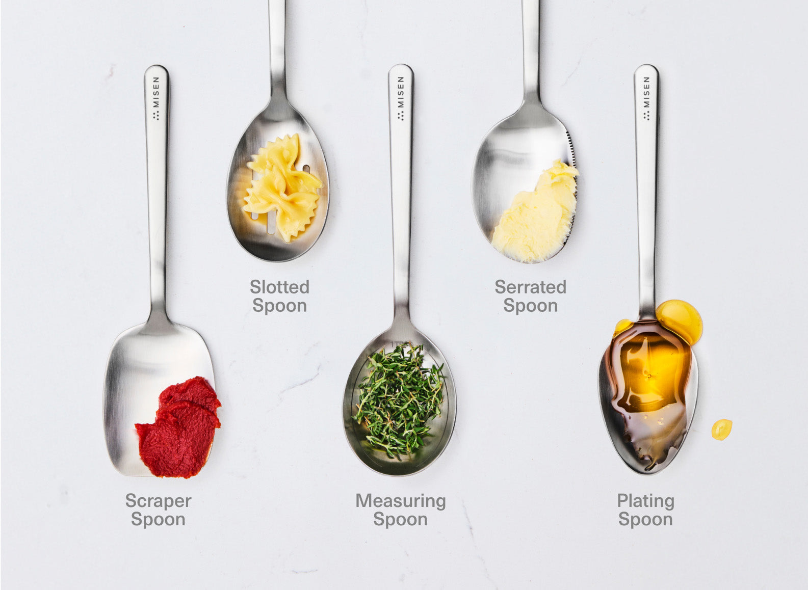 The image displays five stainless steel chef spoons from the brand Misen, each holding a different food item, arranged on a white surface. From left to right, the utensils and their contents are: a scraper chef spoon with tomato paste, a slotted chef spoon with bow-tie pasta, an oval measuring chef spoon with fresh thyme, a serrated-edge chef spoon with butter, and a plating chef spoon with honey. The brand name "MISEN" is embossed on the handle of each utensil.