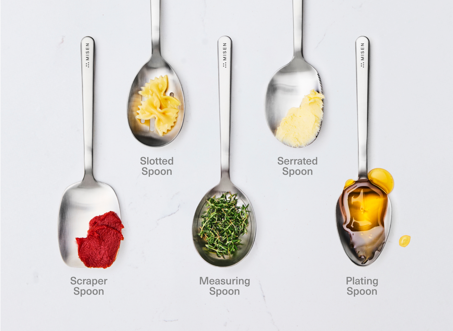 The image displays five stainless steel chef spoons from the brand Misen, each holding a different food item, arranged on a white surface. From left to right, the utensils and their contents are: a scraper chef spoon with tomato paste, a slotted chef spoon with bow-tie pasta, an oval measuring chef spoon with fresh thyme, a serrated-edge chef spoon with butter, and a plating chef spoon with honey. The brand name "MISEN" is embossed on the handle of each utensil.