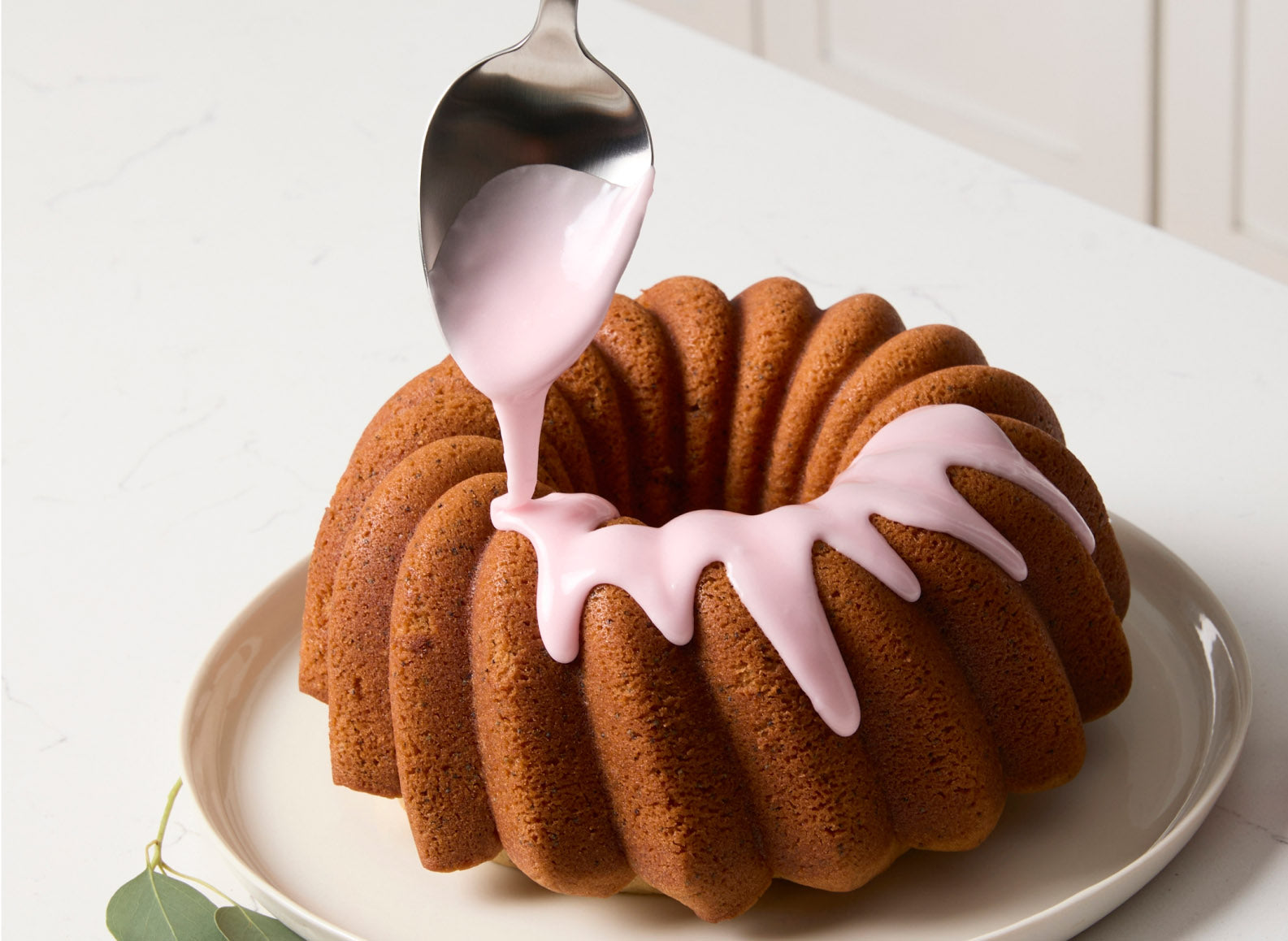 A light pink glaze is being drizzled from a Misen stainless steel plating spoon onto a golden-brown bundt cake. The cake, with its fluted design, rests on a light-colored plate. To the right of the cake, a bunch of red currants with green leaves is visible. To the left, a sprig of greenery with red berries is also visible.