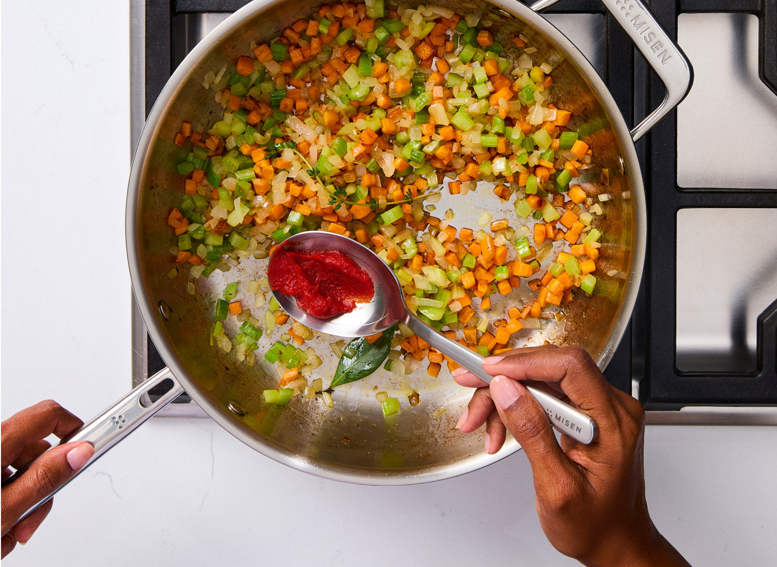 An overhead shot of a woman's hands holding a Misen stainless steel measuring spoon with a dollop of tomato paste, over a large skillet filled with mirepoix. The vegetables, including carrots, celery, and onions, appear to be sautéing. The sauté pan has two handles, one of which is being held by the person. The word "MISEN" is visible on the handle of the spoon and on the handle of the sauté pan.