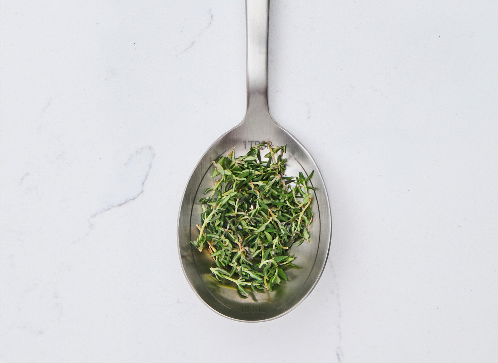 An overhead shot of a stainless steel Misen measuring spoon filled with fresh thyme sprigs. The spoon is resting on a white marble countertop. Engraved measurements, "1 TBSP" and "1 TSP", are visible on the interior of the spoon.