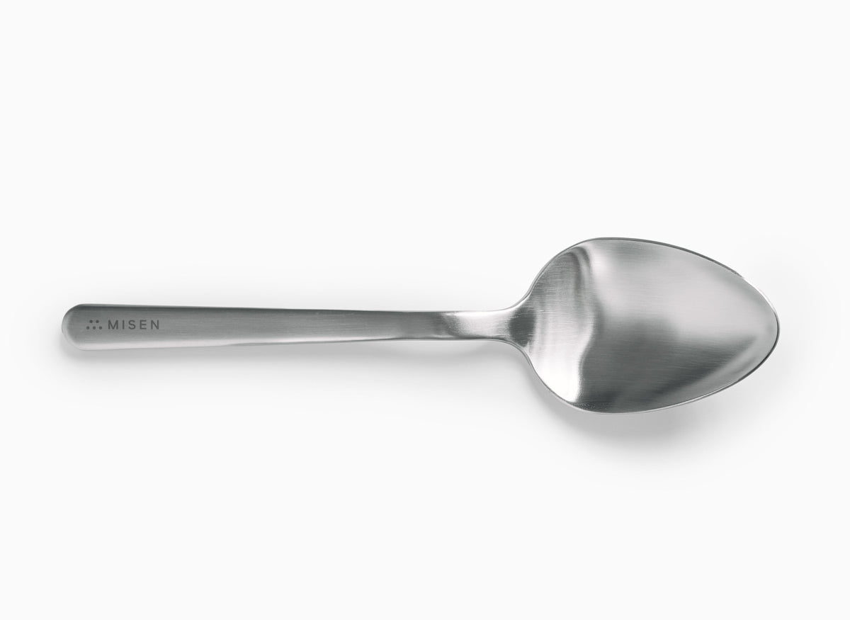 A Misen stainless steel plating spoon is centered horizontally on a white background. It has a polished, reflective oval-shaped bowl and a long, slender handle with a matte finish. The word "MISEN" is debossed on the handle near the end, preceded by a logo of five small dots.