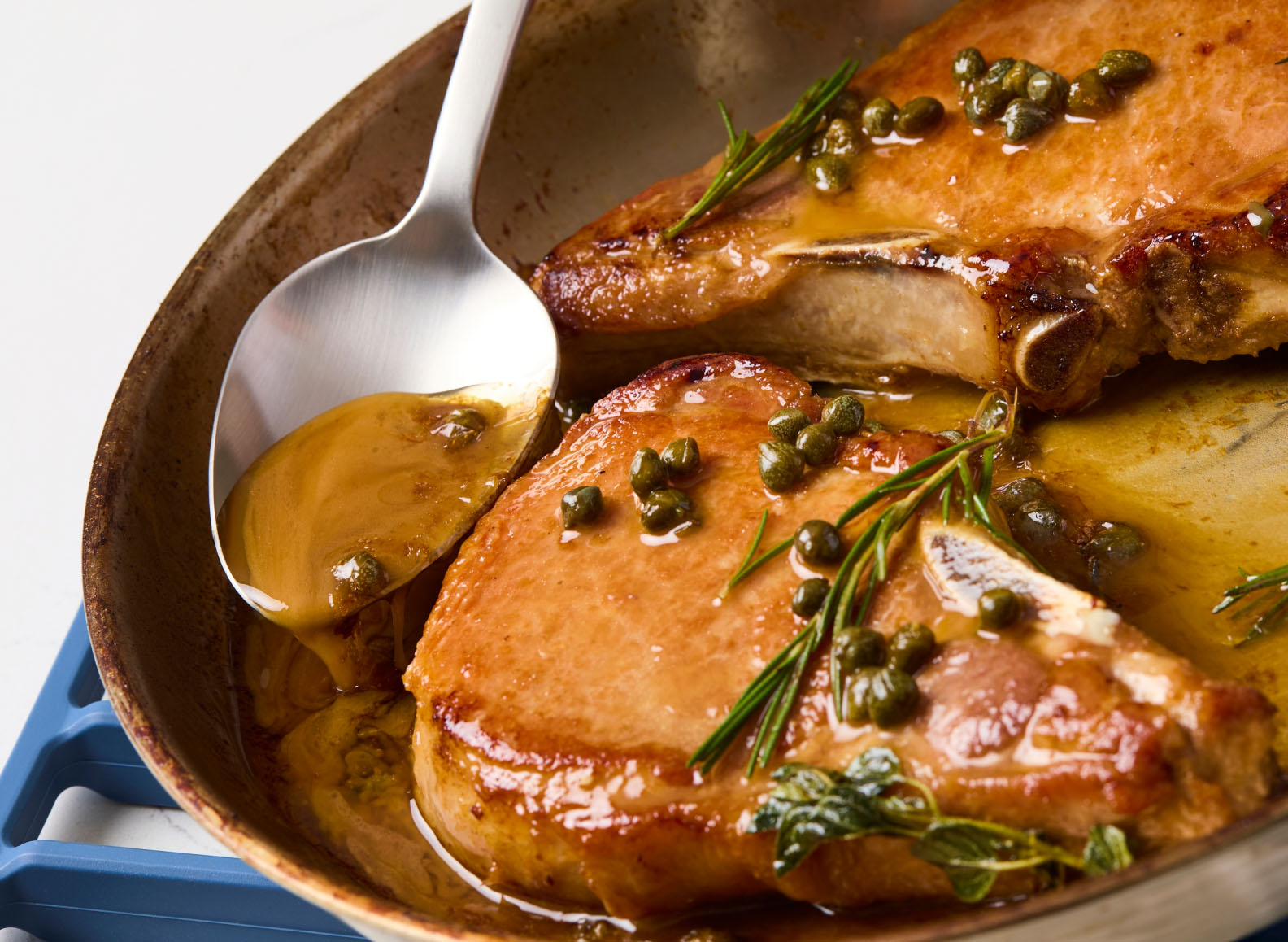 A Misen plating spoon is used to baste two thick, cooked pork chops in a pan. The pork chops are glistening with a rich, golden sauce and garnished with capers and sprigs of fresh rosemary and what appears to be thyme. The pan has a light-colored, rustic-looking exterior and is sitting on a blue Misen silicone roasting rack.