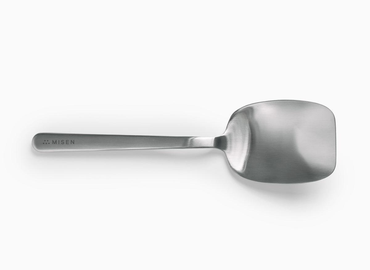 A Misen Scraper Chef Spoon is centered horizontally on a white background. It has a brushed finish and a flat, somewhat rectangular head with rounded corners. The handle is slender and widens slightly where it meets the spoon head. The word "MISEN" is debossed on the handle near the end, with a logo of five small dots preceding it.