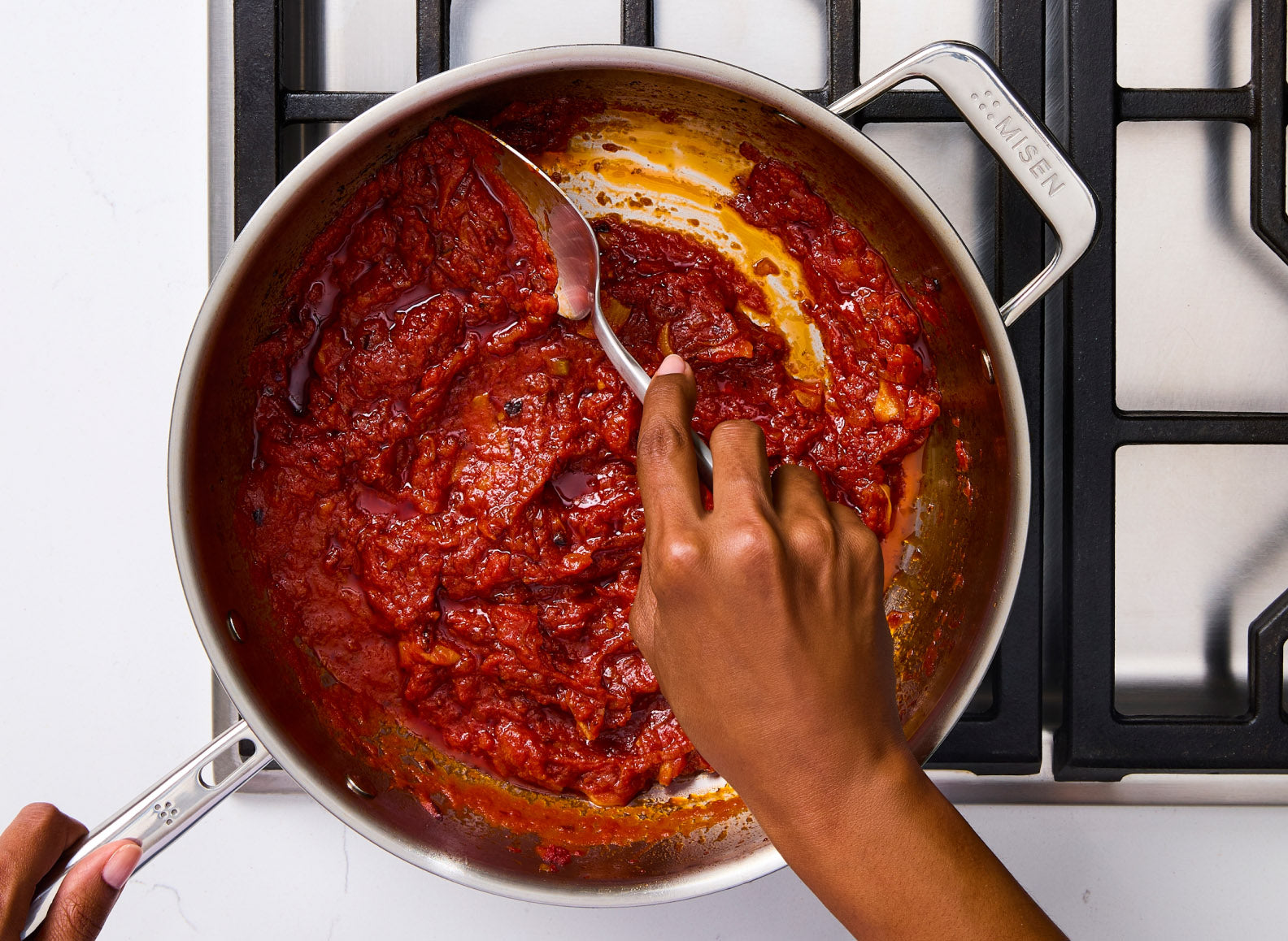 A hand uses a Misen Scraper Chef Spoon to spoon a rich red tomato sauce in a stainless steel sauté pan. The pan has a handle on the right with the word "MISEN" embossed on it.
