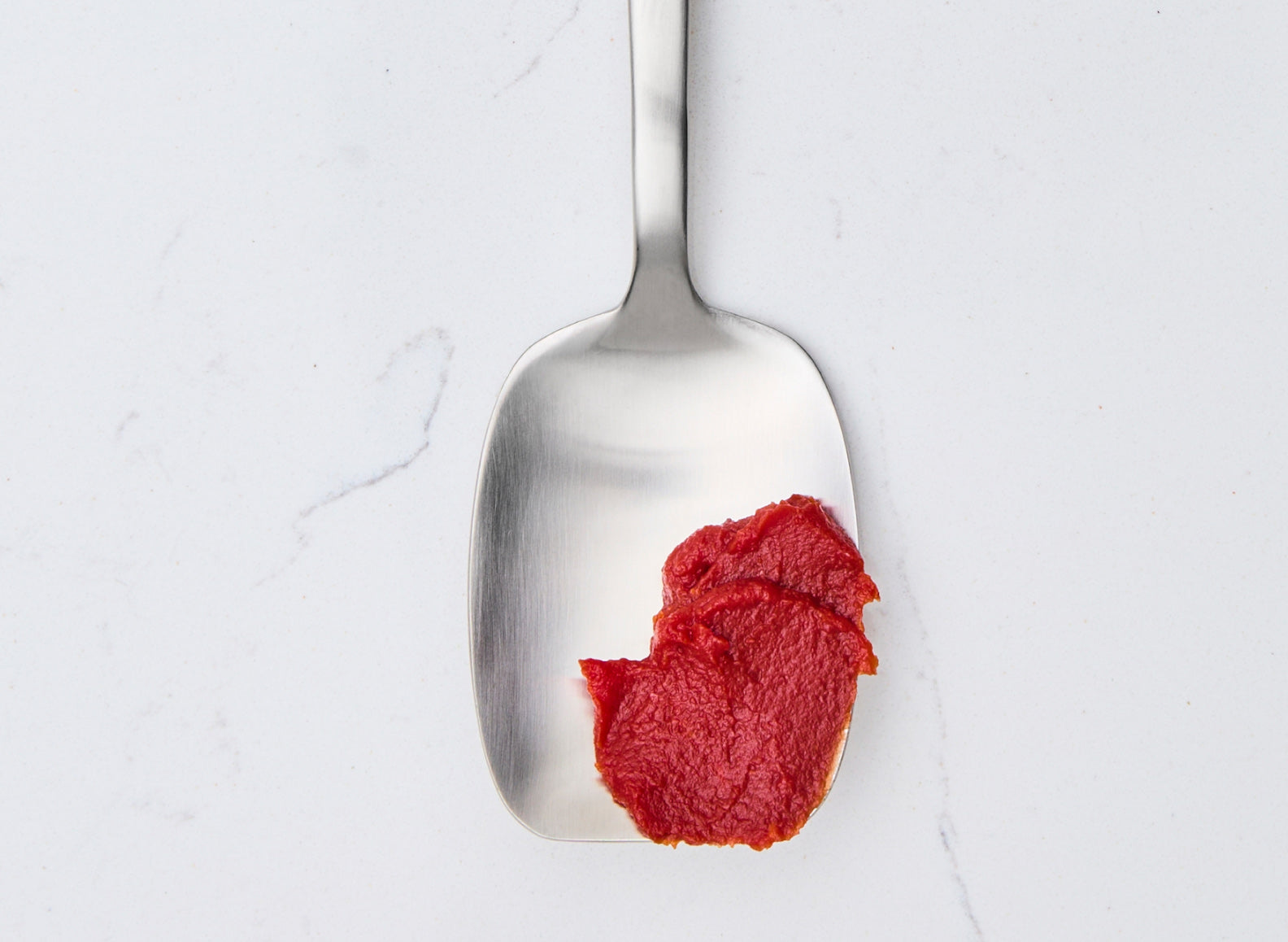 A stainless steel scraper-spoon, with a wide, flat, and somewhat rectangular head, is positioned vertically on a white marble surface. A dollop of thick red tomato paste is on the lower right side of the spoon's head. The spoon has a brushed finish.