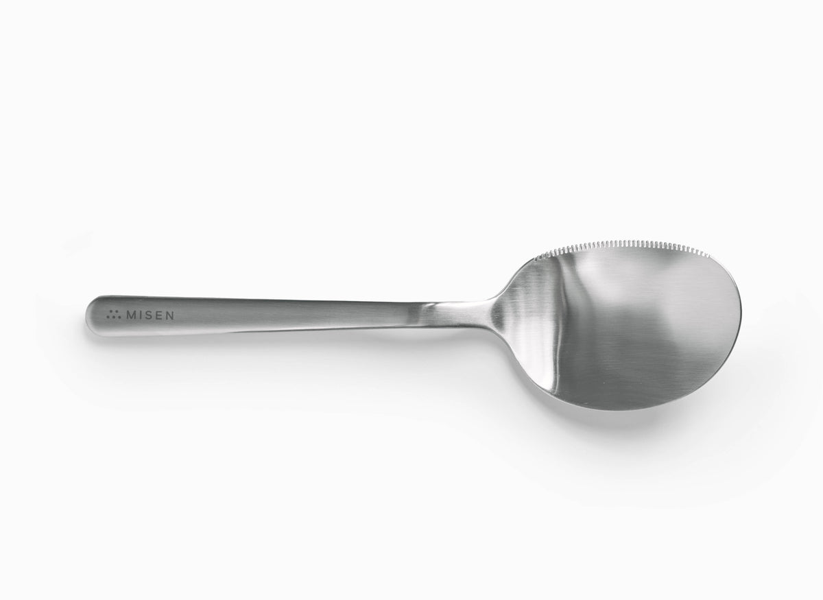 A serrated Misen stainless steel serving spoon is centered horizontally on a white background. It has a polished, reflective oval-shaped bowl with serrations along the left half of its rim. The handle is slender and widens slightly where it meets the spoon head. The word "MISEN" is debossed on the handle near the end, preceded by a logo of five small dots.