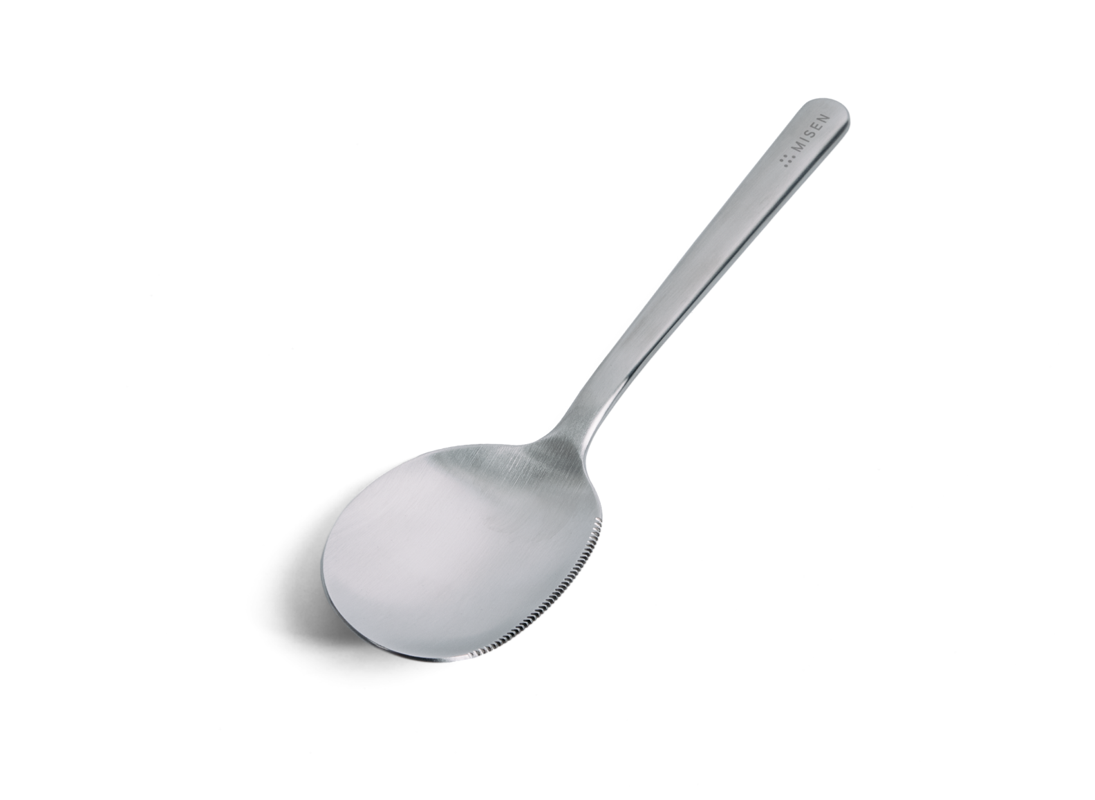A serrated Misen stainless steel serving spoon is positioned diagonally against a white background, with its handle extending towards the upper right. The spoon has a deep, oval-shaped bowl with serrations along the left half of its rim and a matte or brushed finish. The word "MISEN" is subtly debossed on the upper right side of the handle, near its end.