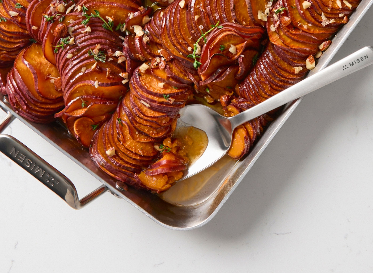 A serrated Misen stainless steel serving spoon is scooping out a portion of thinly sliced, roasted sweet potatoes arranged in an overlapping pattern in a Misen Roasting Pan. The sweet potatoes are glazed and garnished with chopped nuts and fresh thyme sprigs. The roasting pan has handles on either side, one of which reads "MISEN".