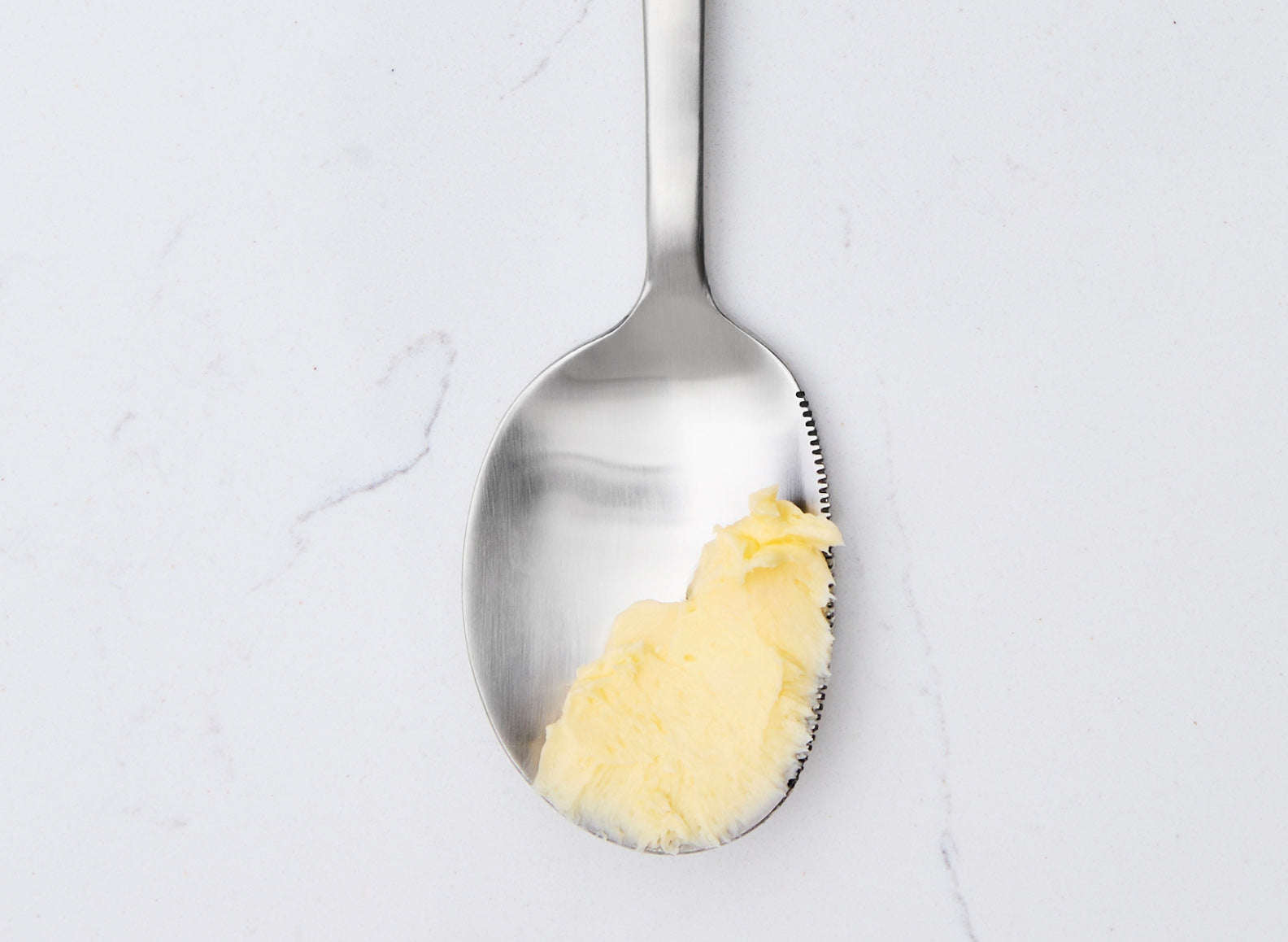 A serrated Misen stainless steel serving spoon holds a scoop of yellow butter. The spoon is positioned vertically on a white marble countertop, with its serrated edge facing right. The butter is soft and unevenly shaped.