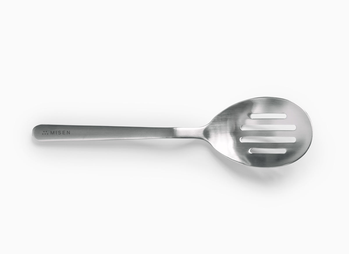A Misen stainless steel slotted spoon is positioned horizontally on a white background. The spoon has an oval-shaped head with four horizontal slots for drainage. The handle is slender and widens slightly where it meets the spoon head. The word "MISEN" is debossed on the handle near the end, preceded by a logo of five small dots.
