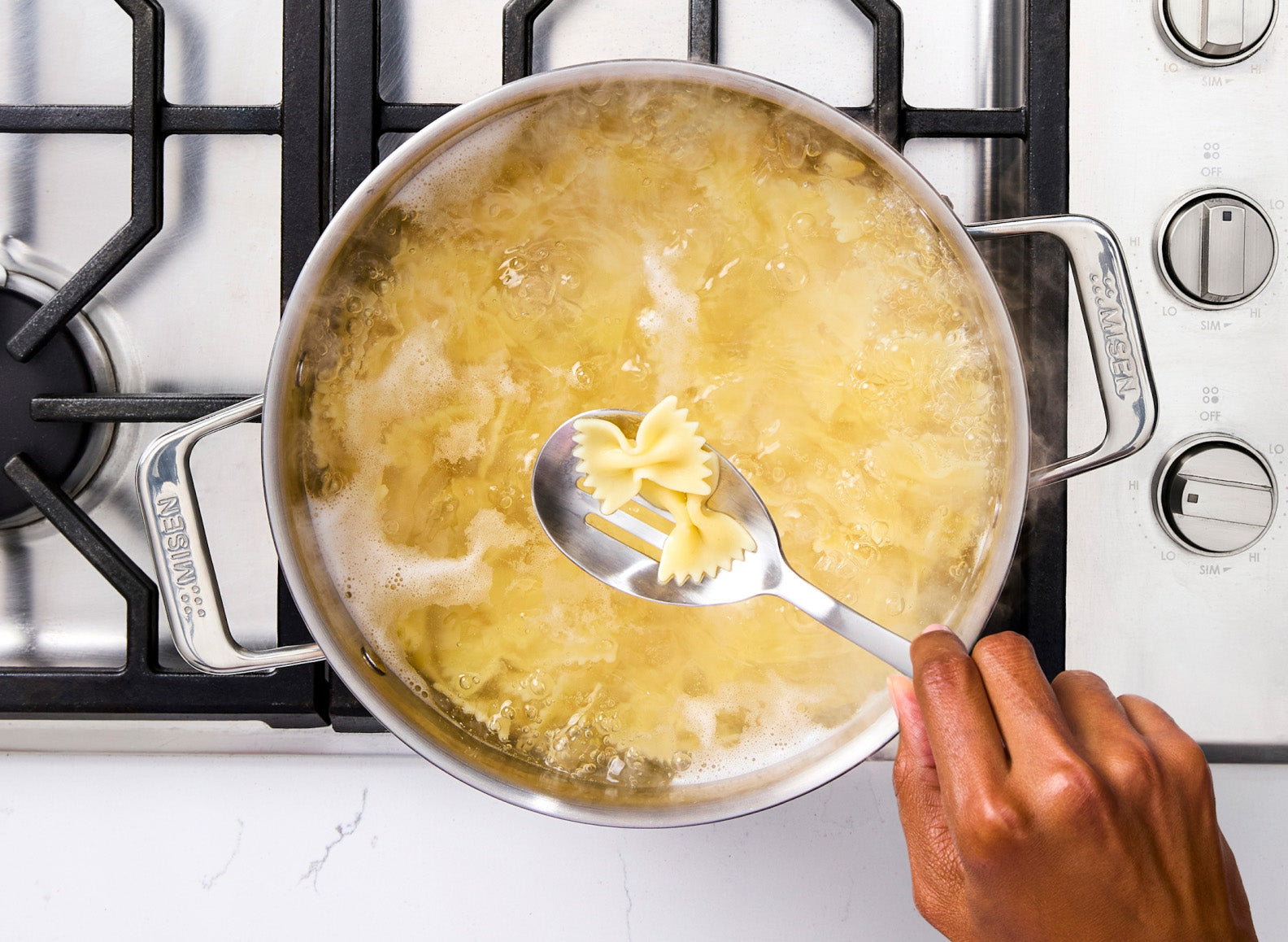 A hand holds a stainless steel slotted spoon with a few bow-tie pasta pieces, lifting them from a large stainless steel rondeau of boiling water on a gas stovetop. The rondeau has handles on either side, each embossed with "MISEN". The stovetop burners and control knobs are visible around the pot.