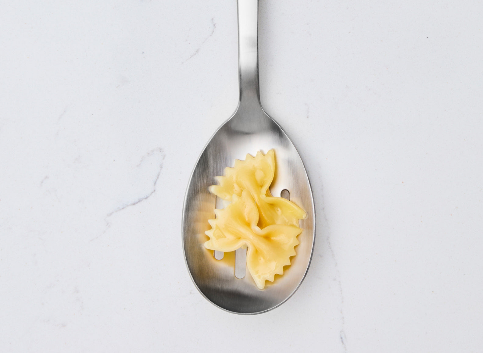 A Misen stainless steel slotted spoon holds two pieces of cooked bow-tie pasta. The spoon is centered vertically against a white marble countertop. The pasta is light yellow.
