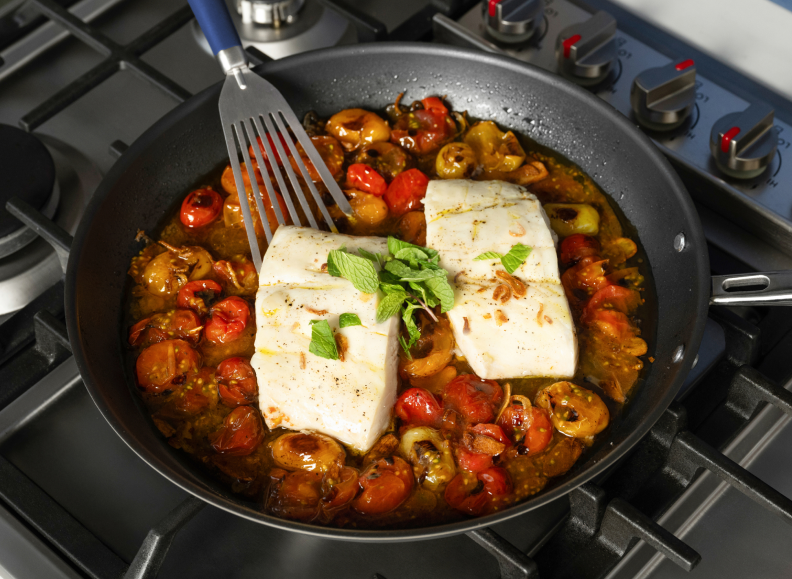 Two seasoned fillets of white fish and cherry tomatoes are cooking in a Misen Carbon Nonstick™ Frying Pan on a stovetop, with a Misen Stainless Steel  Fish Spatula lifting the fish from the pan.