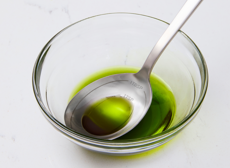 A close-up view shows a stainless steel Misen Measuring Chef Spoon resting in a clear glass bowl containing a green olive oil. The spoon is etched with measurements for 1 tbsp and 1 tsp.