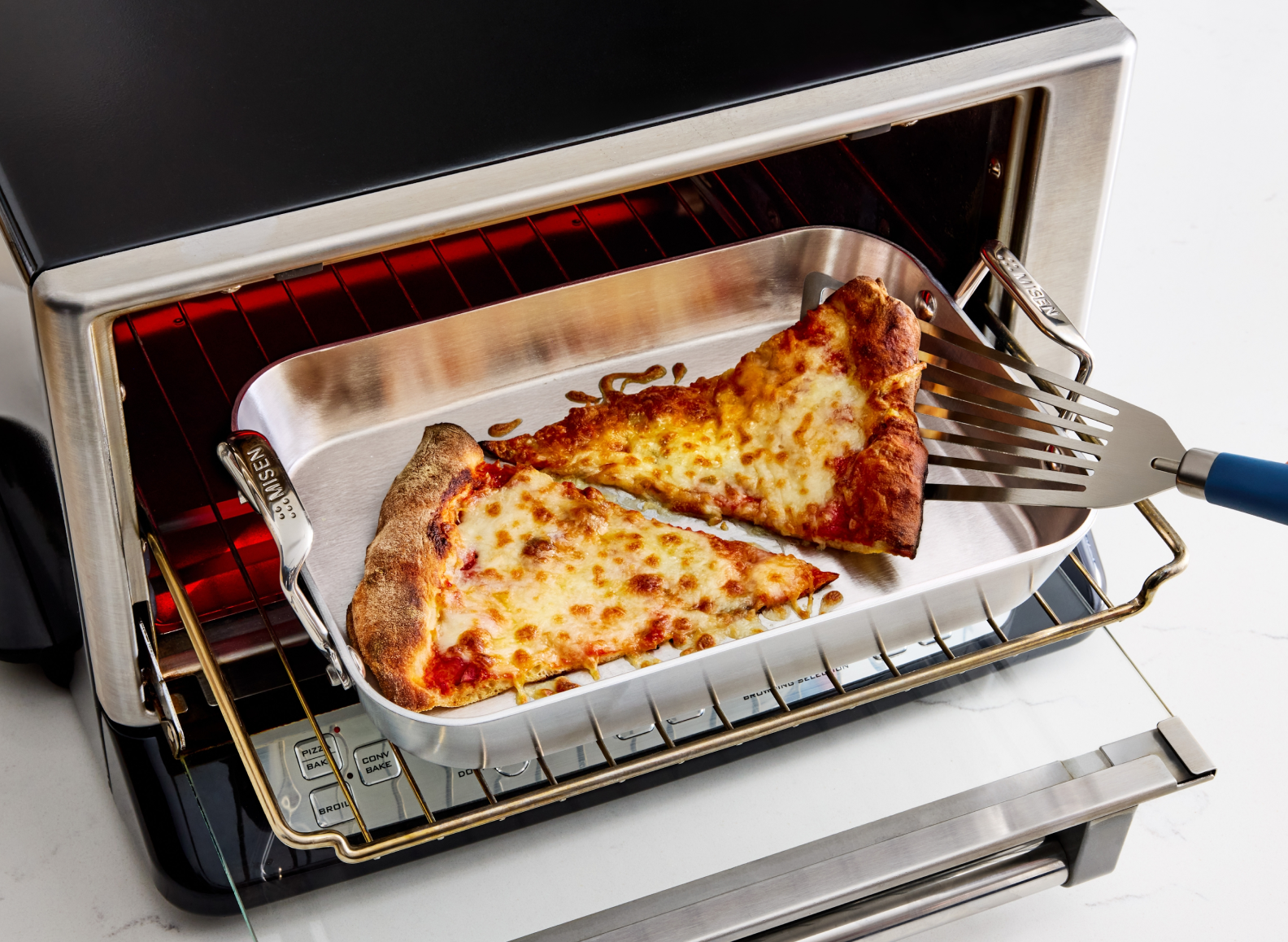 A close-up shot of two slices of cheese pizza being reheated in a small, 9 inch stainless steel roasting pan, visible inside a toaster oven with the door partially open. A stainless steel fish spatula with a blue handle is reaching into the oven to lift one of the slices. The word "MISEN" is visible on the side handles of the pan. The interior of the oven shows the glowing red heating elements.