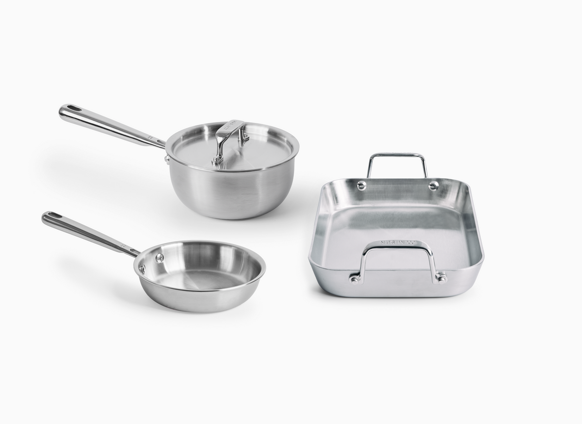 A studio shot on a white background displays three stainless steel Misen cookware items. In the center is a saucier with a long handle and a lid. In the lower left is a small stainless steel frying pan with a long handle. To the right is a small, roasting pan with two raised loop handles. The word "MISEN" is visible on the handles of the frying pan and the roasting pan.