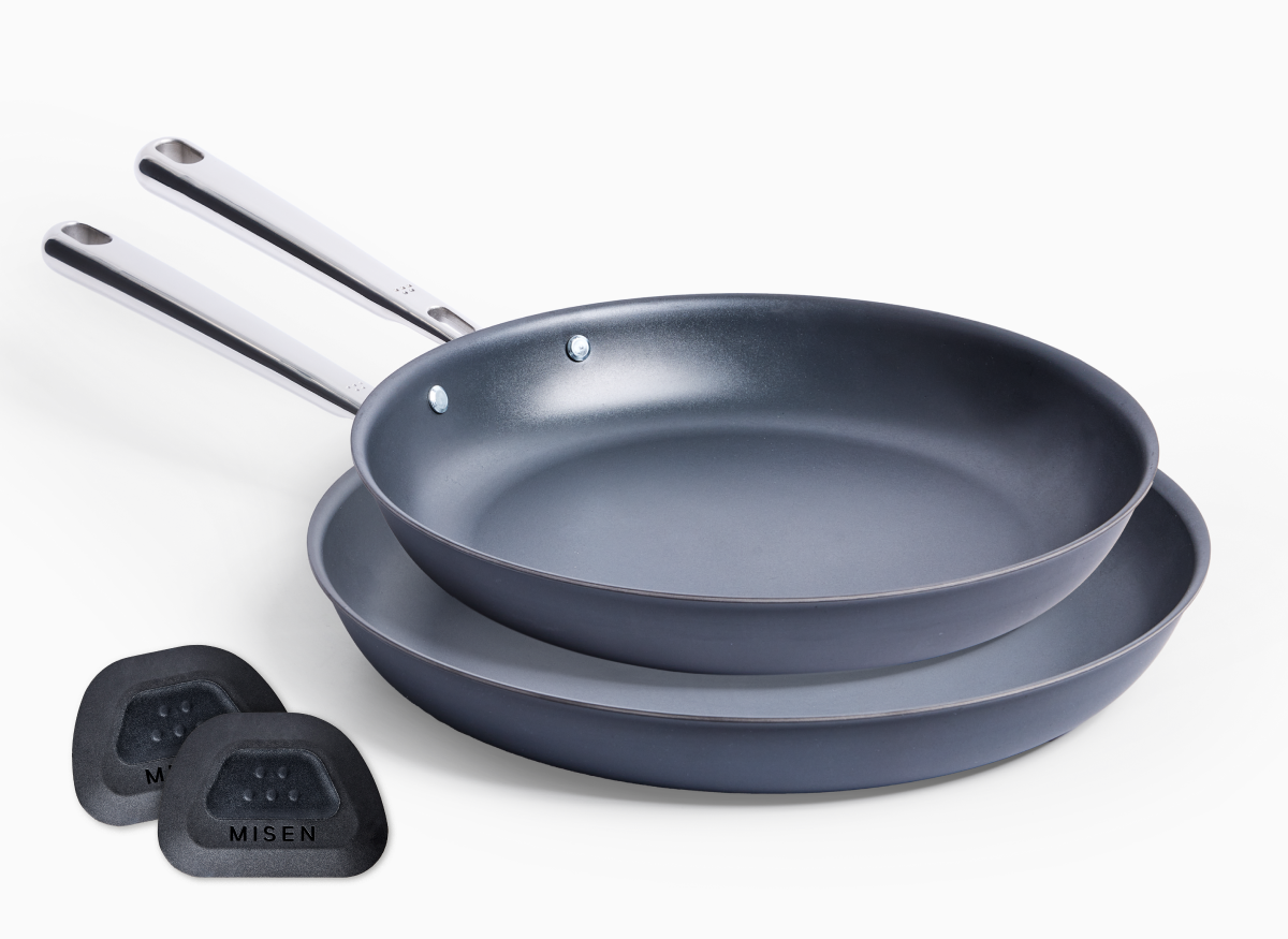 A studio shot on a white background shows a stack of two Misen Carbon Nonstick™ frying pans in a dark charcoal gray color. The top 10 inch pan is slightly smaller and resting inside the bottom 12 inch pan. Both pans have long, polished stainless steel handles. To the bottom left of the stack are two black pan scrapers, with the word "MISEN" visible.