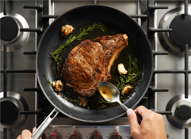 A close-up overhead shot of a thick cut of meat, a steak, searing in a Misen Carbon Nonstick™ Frying Pan on a gas stove. A sprig of rosemary and several cloves of garlic are in the pan with the steak, and a hand holds a spoon, basting the meat.