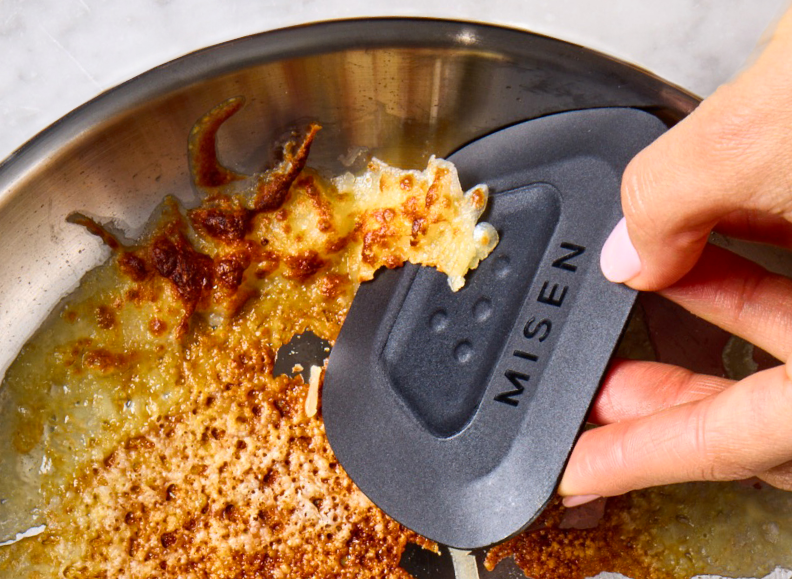 A person's hand uses a black Misen Pan Scraper to remove burnt food from the bottom of a stainless steel saucier. The saucier contains a large amount of burnt, golden-brown cheese.