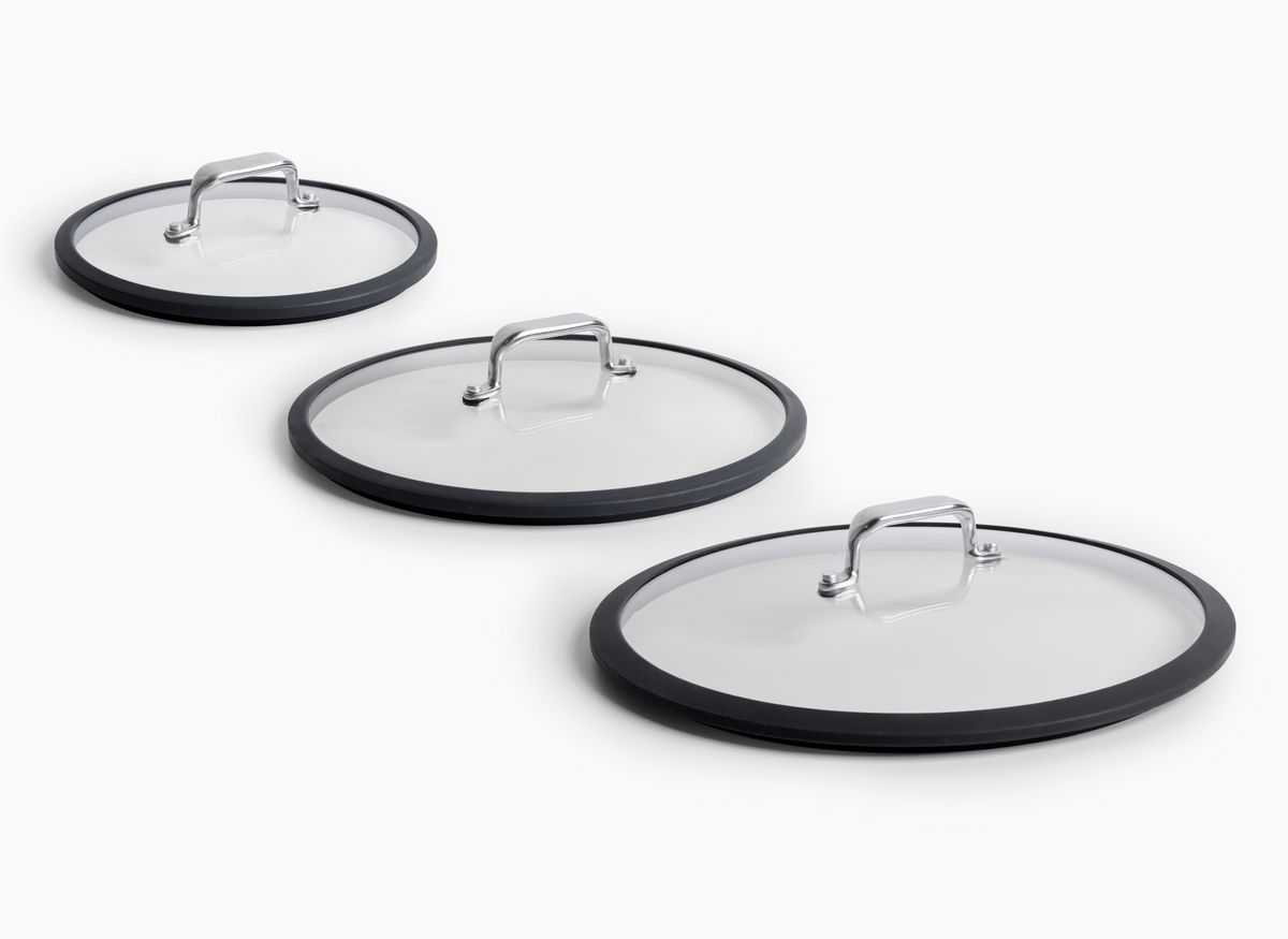 Three round Misen Glass Lids are arranged diagonally on a white surface, with the largest 12 inch lid in the foreground, the smallest 8 inch lid in the background, and the 10 inch lid in the middle. Each lid has a transparent glass center, a black silicone rim, and a silver-colored handle.