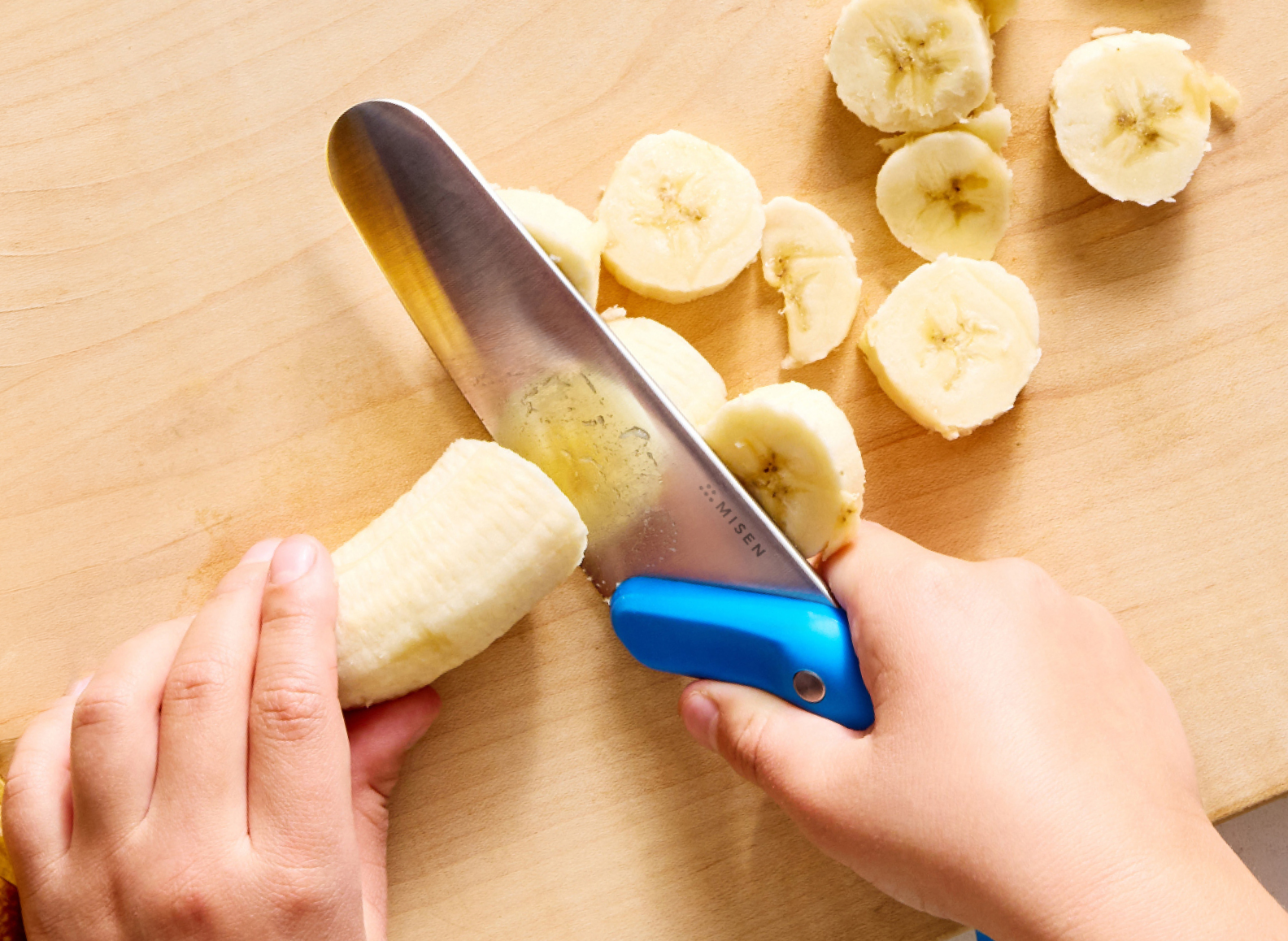 A close-up image showing a child's hands using a blue-handled Misen Kids' Knife to slice a peeled banana on a light wooden cutting board. Several slices of banana are scattered on the board. The word "MISEN" is visible in small print on the side of the knife blade.