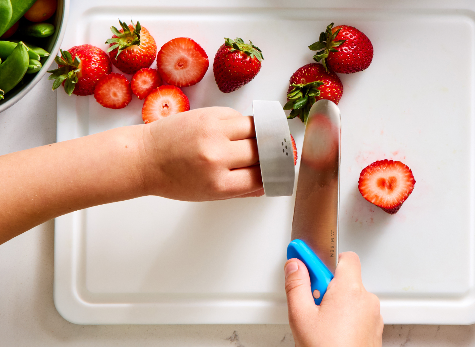 An overhead shot shows a child's hands chopping strawberries on a white cutting board. The right hand holds a Misen kids' knife with a blue handle and a stainless steel blade. The left hand wears a stainless steel finger guard while holding the strawberries steady. Several whole and sliced strawberries are visible on the cutting board. A metal bowl containing produce is partially visible in the upper left corner. The word "MISEN" is visible on the knife blade.