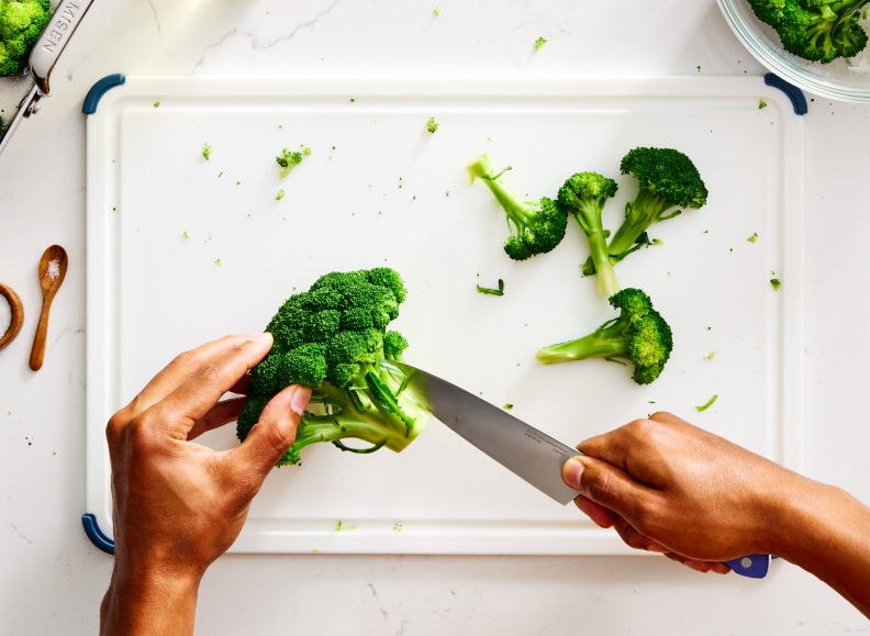 A person's hands are shown from a top-down perspective, cutting a head of broccoli into florets on a white cutting board with a blue-handled Misen Utility Knife 2.0. Several cut florets are scattered across the board, and a small wooden spoon with salt is visible in the upper left corner.