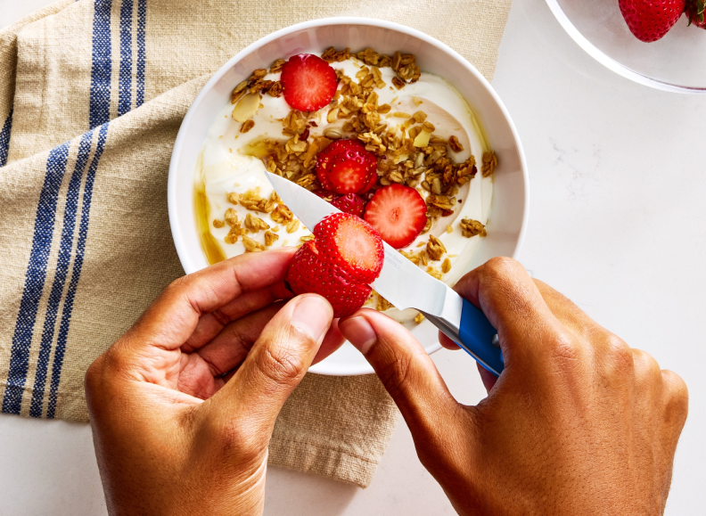 A person's hands are shown from above, slicing a strawberry with a blue Misen 3.5 inch Paring Knife 2.0 over a white bowl filled with yogurt, granola, sliced almonds, and strawberries. A blue striped Misen Kitchen Towel is to the left of the bowl, and a clear bowl with whole strawberries is visible in the upper right corner.