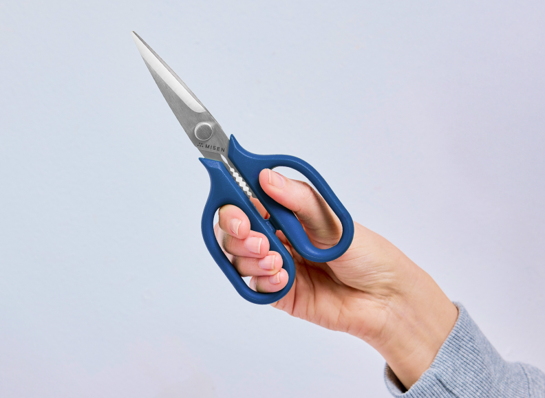 A person's hand holds a blue-handled pair of small Misen Kitchen Shears, with the blades pointing upwards and slightly to the left. The Misen logo is visible near the pivot screw of the shears.