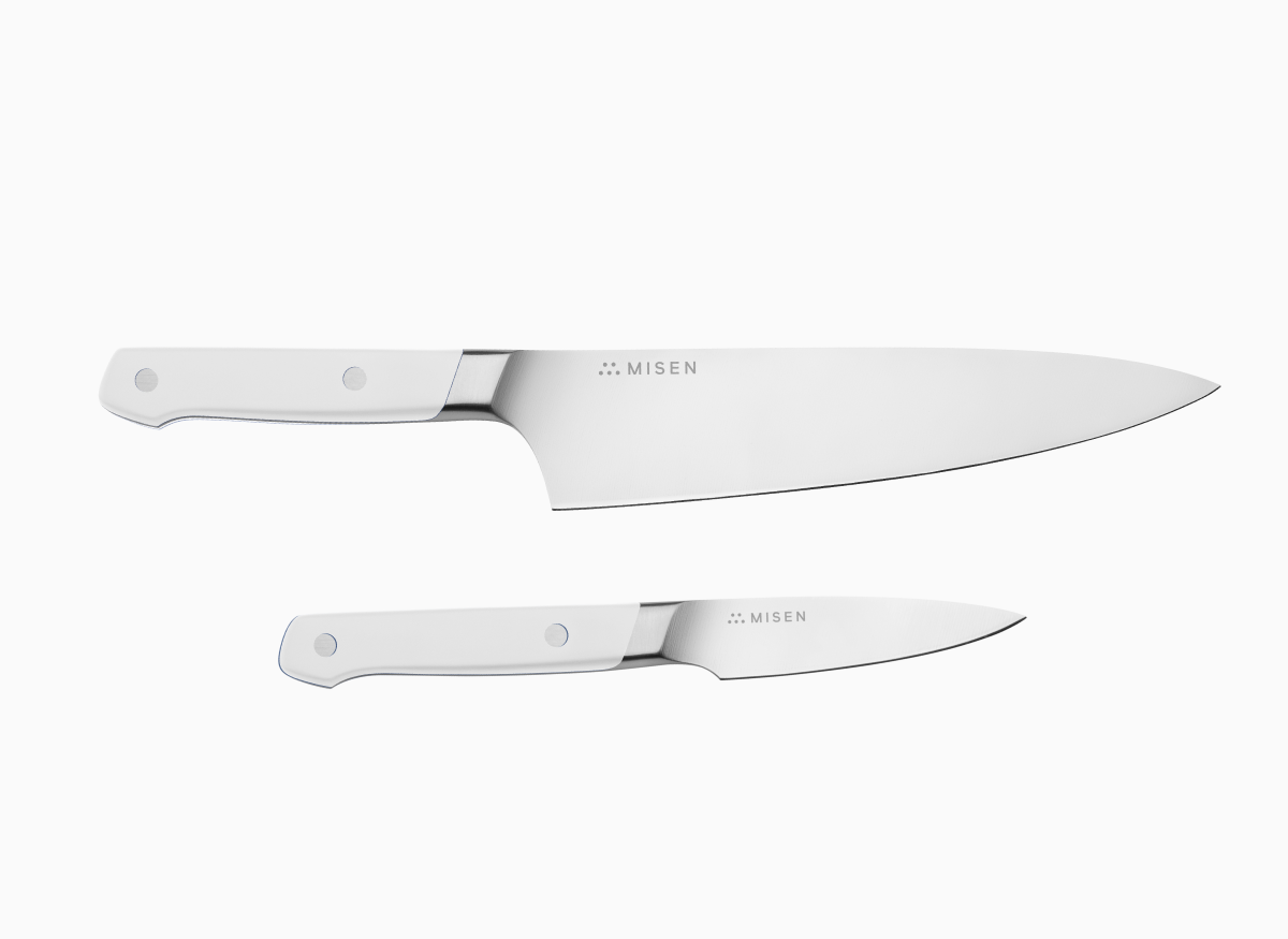 A set of two kitchen knives with white handles and silver blades, one Chef's, one Paring, laid on a white surface.