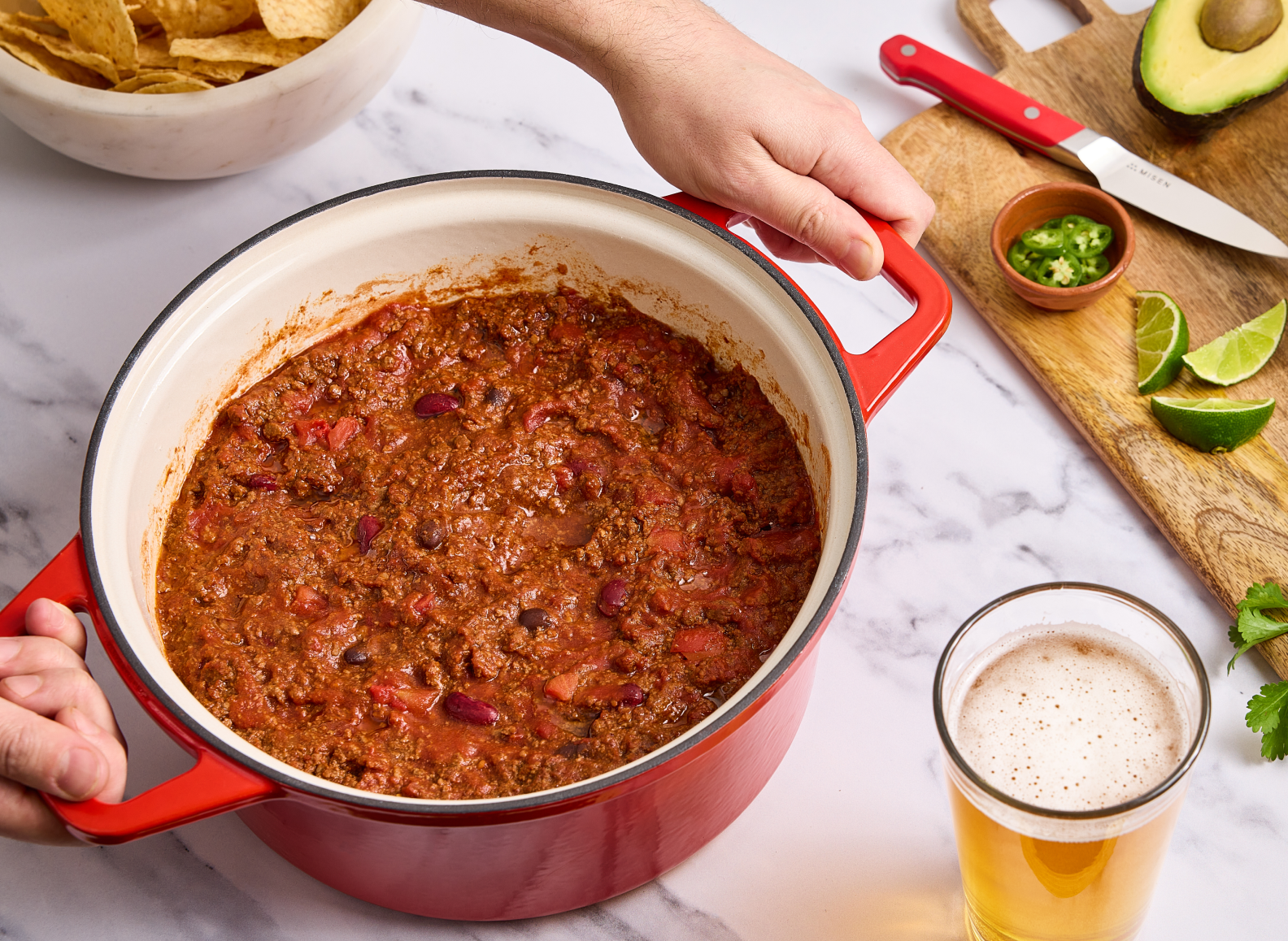 A close-up overhead shot shows a person holding a large red Dutch oven filled with chili. To the left is a white bowl of tortilla chips. To the right, on a wooden cutting board, are a red-handled knife, a small bowl of sliced green chilis, and lime wedges. In the bottom right corner, a glass of beer is visible.