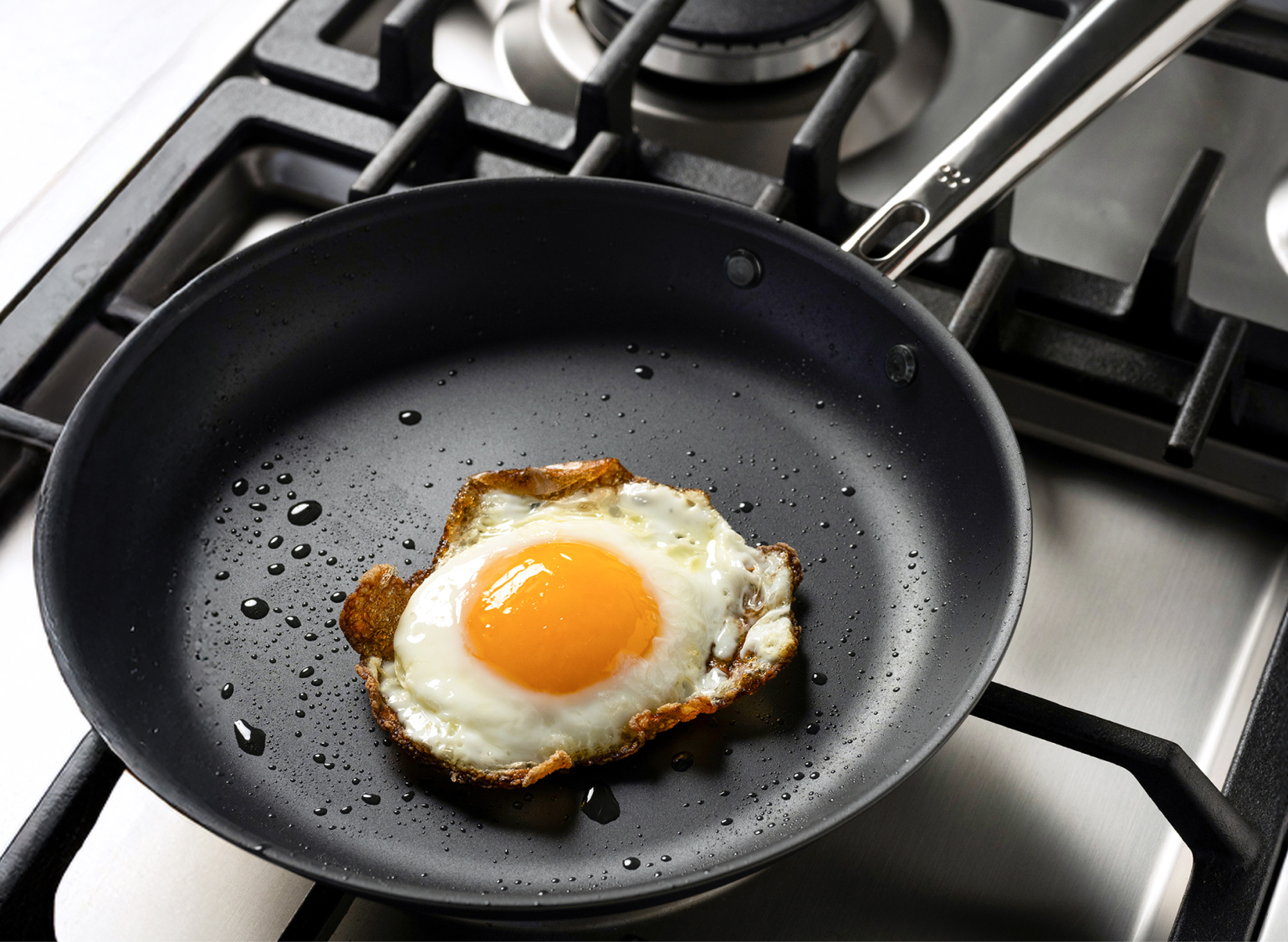 A sunny-side up egg cooking in a Misen Carbon Nonstick™ Frying Pan on a gas stove. The egg white is crispy around the edges, and small droplets of oil are visible in the pan.