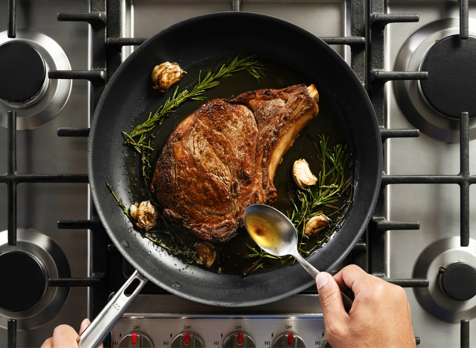 A close-up overhead shot of a thick cut of meat, a steak, searing in a Misen Carbon Nonstick™ Frying Pan on a gas stove. A sprig of rosemary and several cloves of garlic are in the pan with the steak, and a hand holds a spoon, basting the meat.