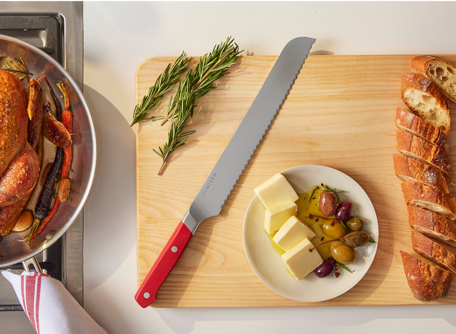 A 10 inch Serrated Knife in red on a wooden cutting board, next to some chopped rosemary, sliced bread, and a bowl of olives marinated with olive oil and cubed butter for dipping & spreading.