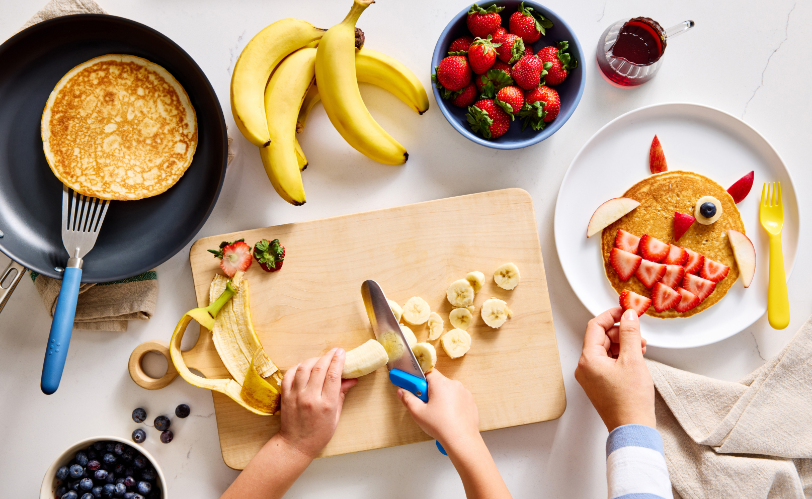 An overhead shot of a breakfast scene. In the center, a child's hands are using a blue-handled Kid's Knife to slice a peeled banana on a light wooden cutting board. To the left, a Carbon Nonstick™ pan contains a golden-brown pancake, with a blue, stainless steel fish spatula resting on the edge of the pan. A bunch of bananas is above the cutting board, and a blue bowl of strawberries is in the upper right. An adult's hands are arranging strawberry slices on a pancake shaped like a monster face.