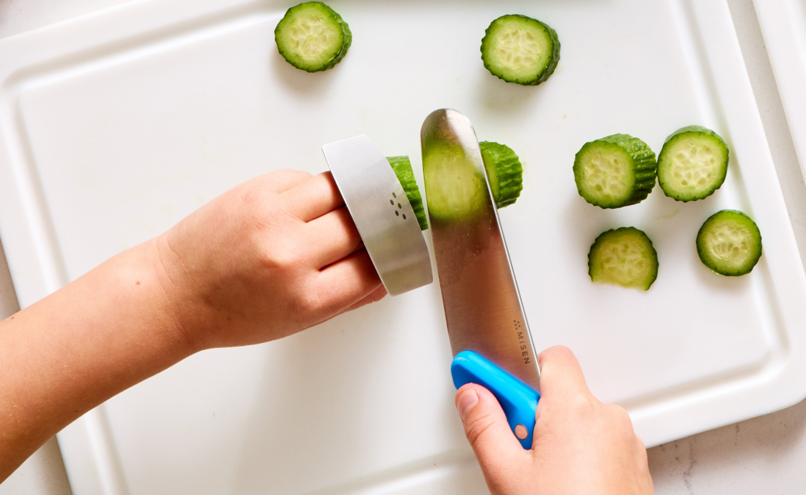 An overhead shot showing a child's hands slicing mini cucumbers on a white cutting board. The right hand holds a Misen Kids' Knife with a blue handle, and the left hand wears a stainless steel finger guard while holding the cucumber steady. Several slices of cucumber are scattered on the cutting board. The word "MISEN" is visible on the knife blade.